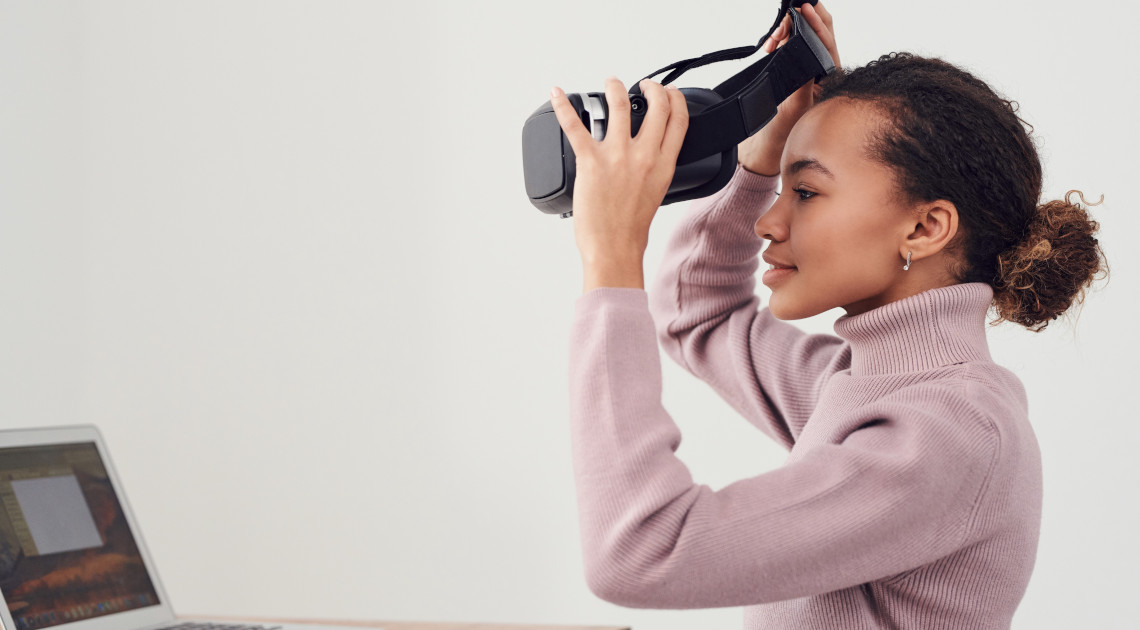Fit für den digitalen Wandel: Eine junge Frau setzt vor einem Laptop ein schwarzes Virtual-Reality-Headset auf (© Pexels/Fauxels)